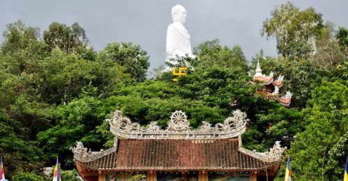 A large statue of Buddha behind a temple and tall trees at the Long Son Pagoda